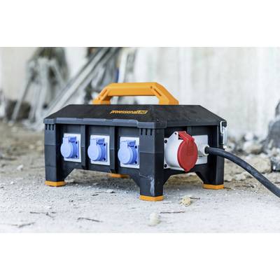 A portable construction site distribution box with multiple blue and one red socket stands on the ground of a construction site.