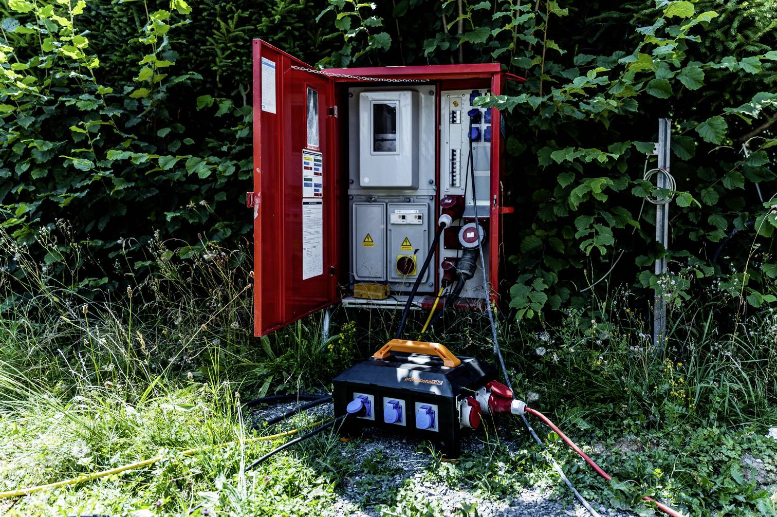An open, red electrical cabinet outdoors, surrounded by plants. An electrical device is connected to the cabinet.