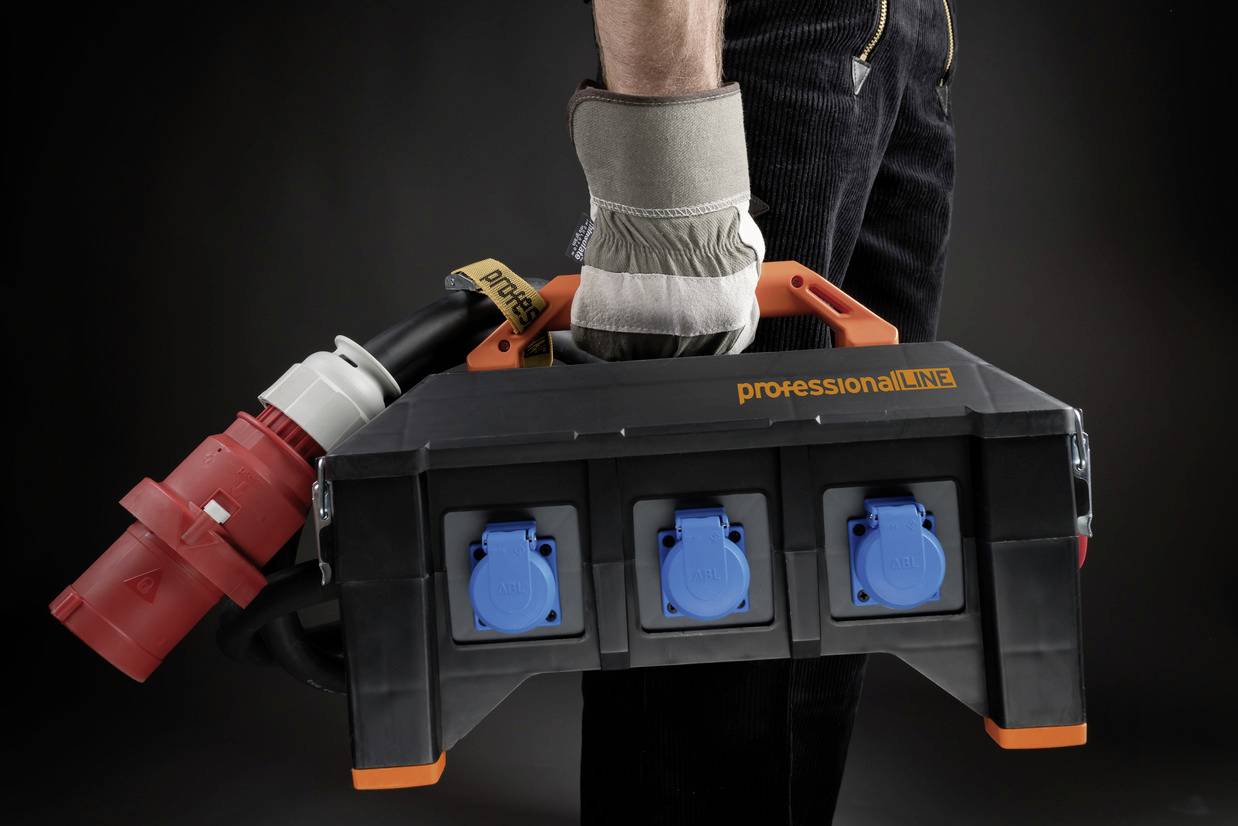 A person is holding a large black cable reel with blue sockets and a thick red plug, accompanied by a grey glove.