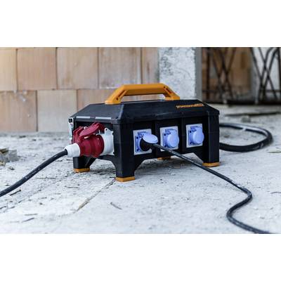 Black and orange power distributor on concrete floor with multiple sockets and connected cables, with raw brick wall in the background.