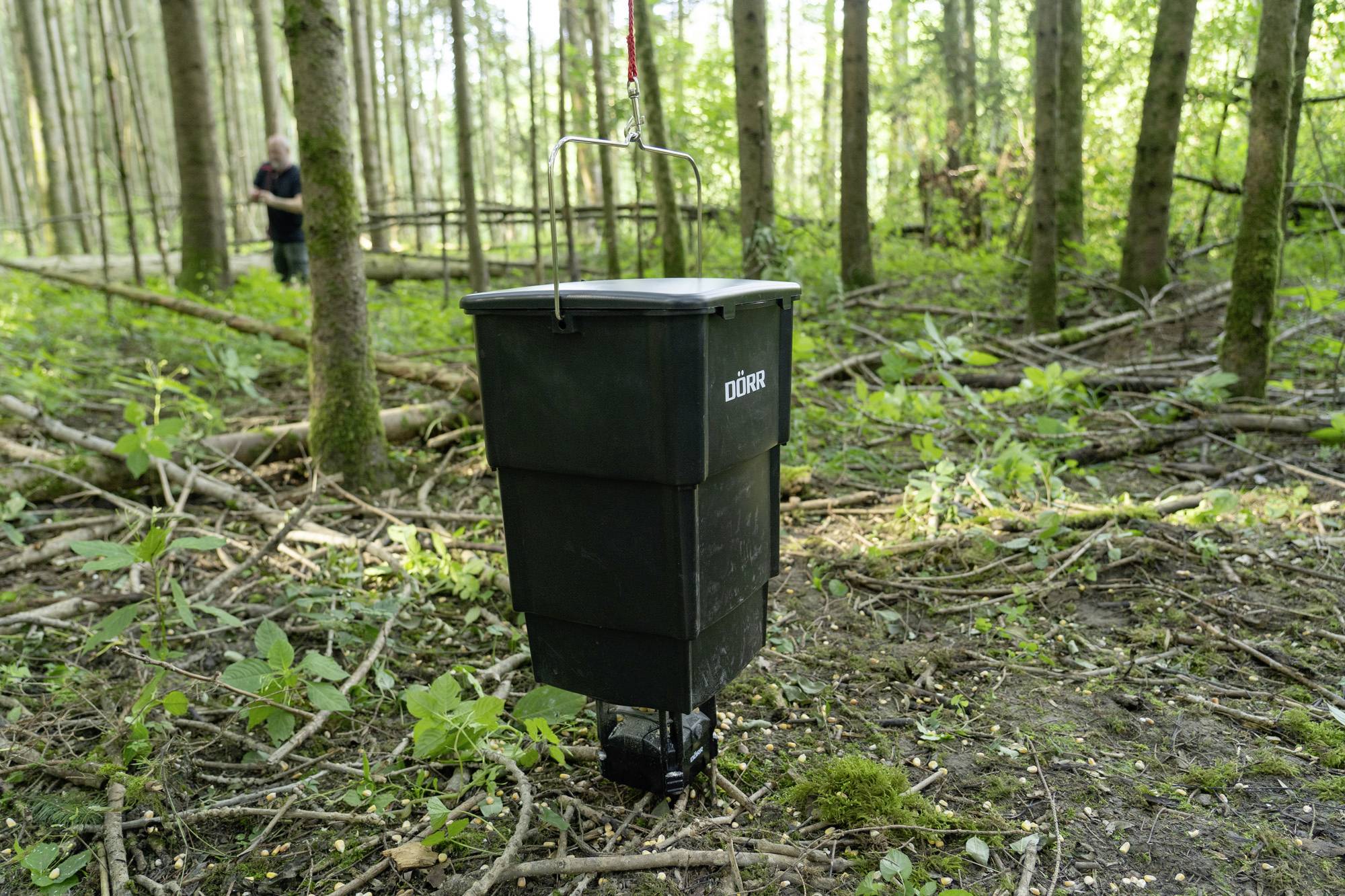 A black feed dispenser is standing on the ground in the forest. A person can be seen blurrily in the background.