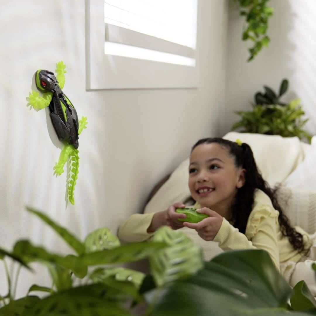 A child plays happily on a sofa with a toy gecko sticking to the wall. Plants can be seen in the background.