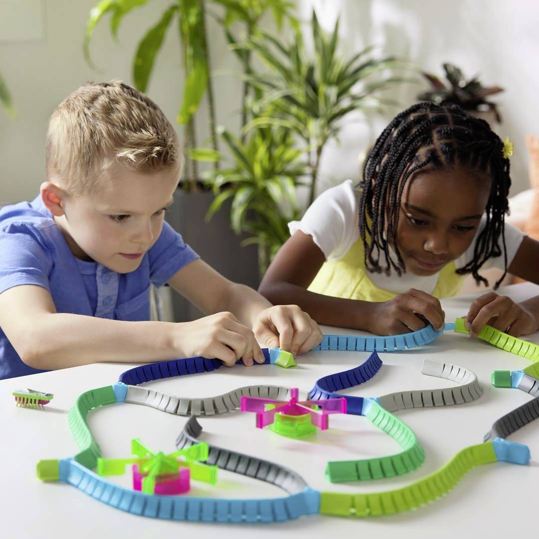 Two children are intently playing with a colourful domino chain set on a table, surrounded by plants in the background.