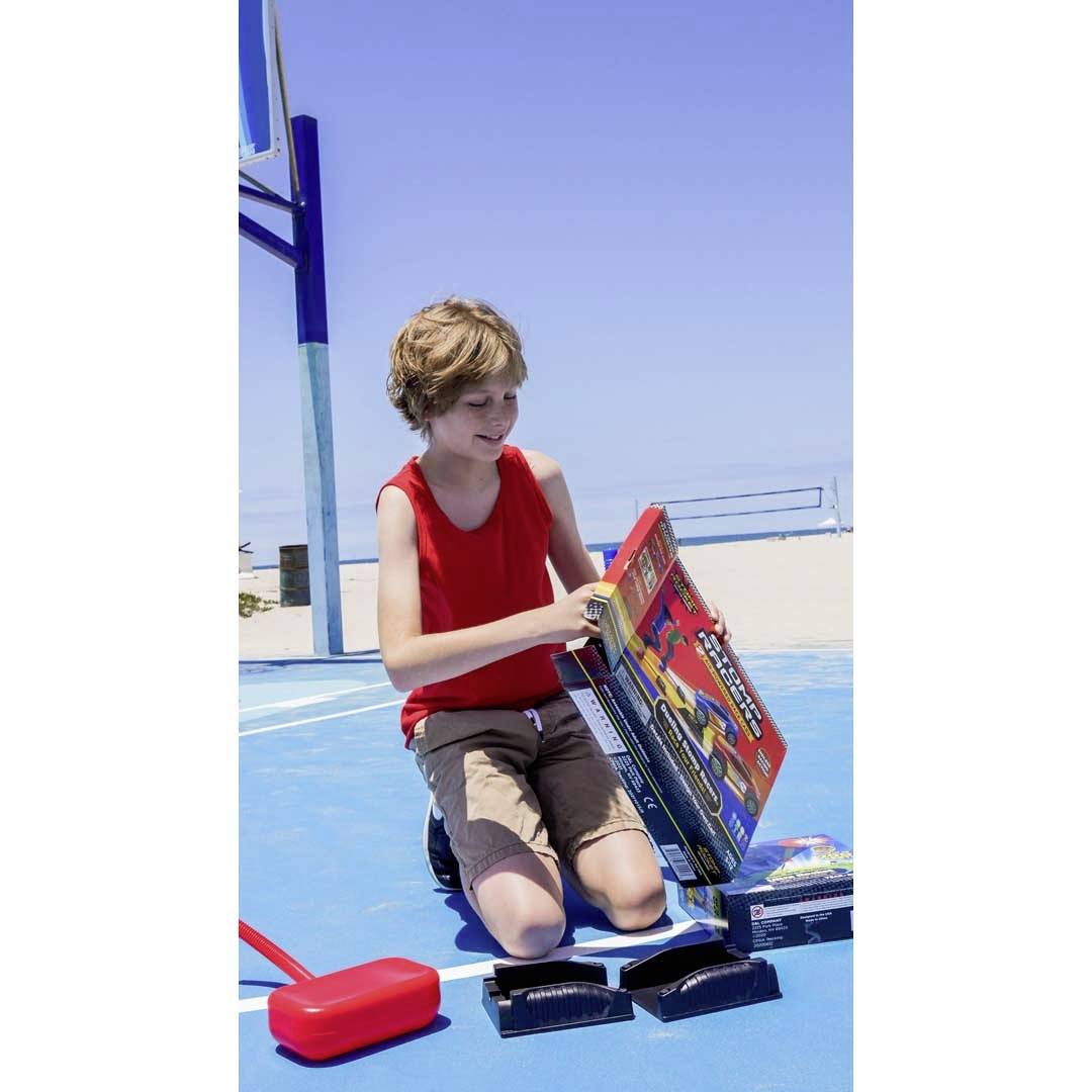 A boy in a red vest kneels on an outdoor blue basketball court and opens a box with a toy racing track.