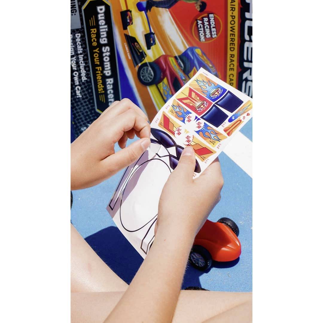 A child is holding stickers and a small toy car with an orange bodywork, with a toy car packaging lying in the background.