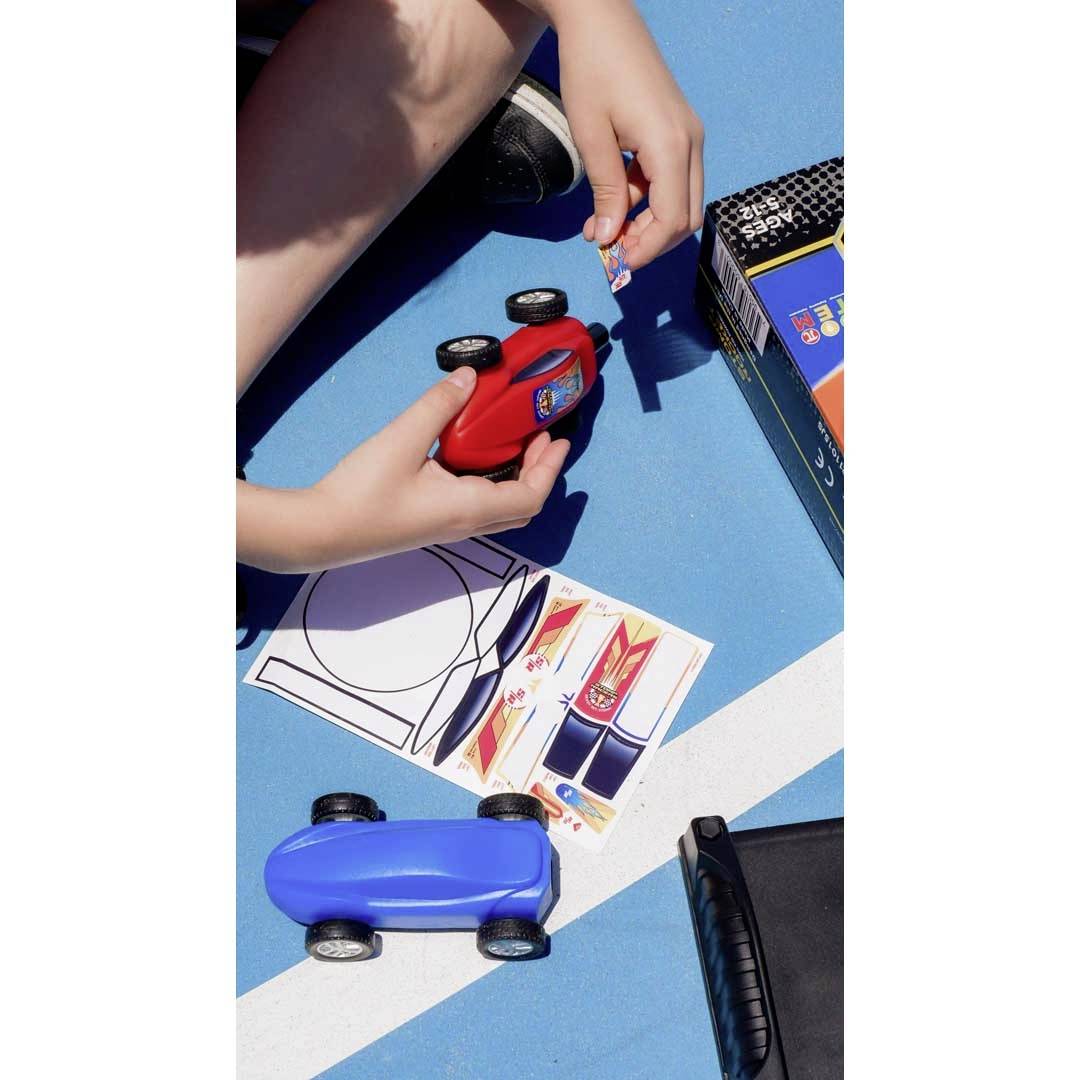 A child on a blue background is sticking stickers onto a red toy car. A blue car and sheets of stickers are lying nearby.
