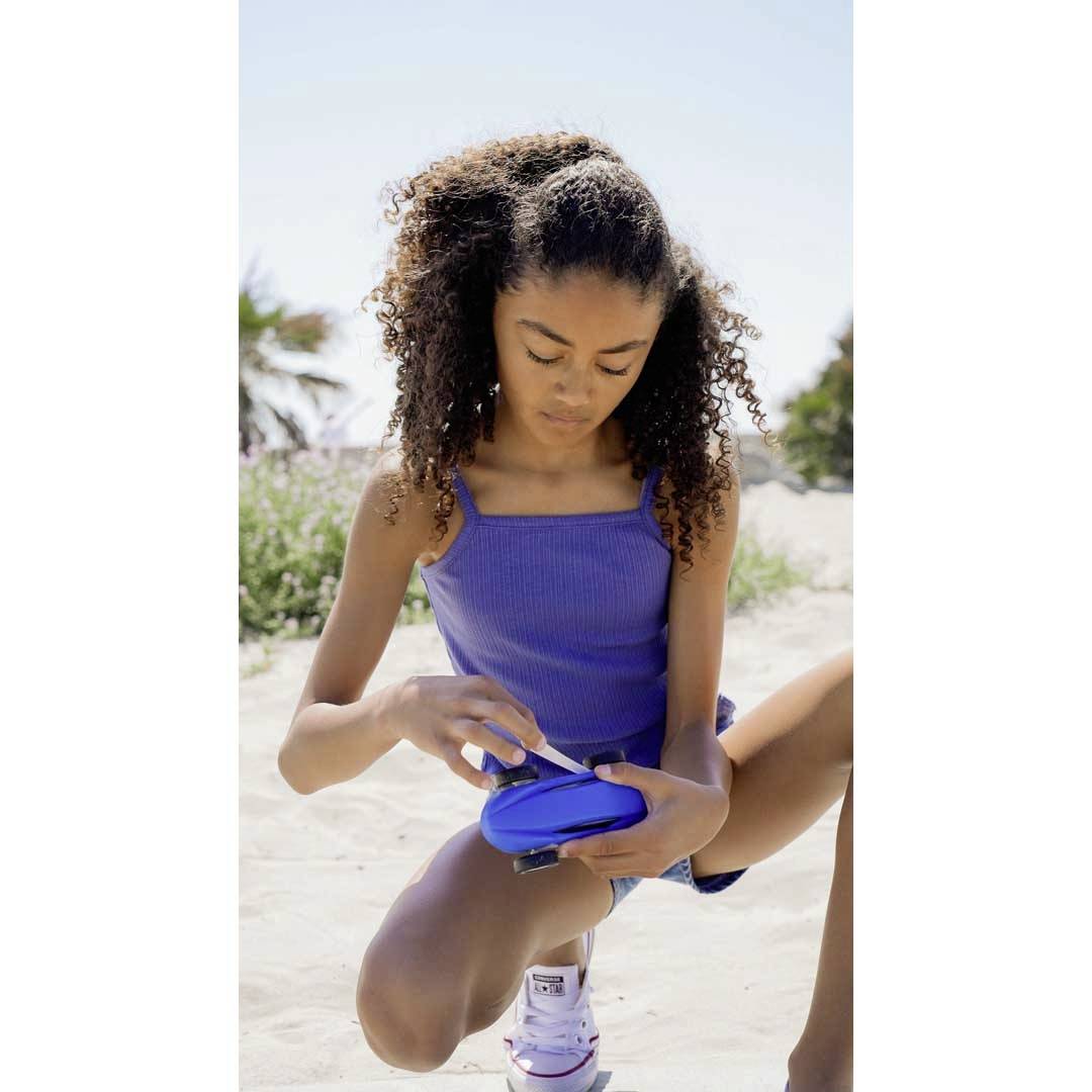 Girl on the beach, kneeling in the sand, taking something from a blue container; palm trees and trees are visible in the background.