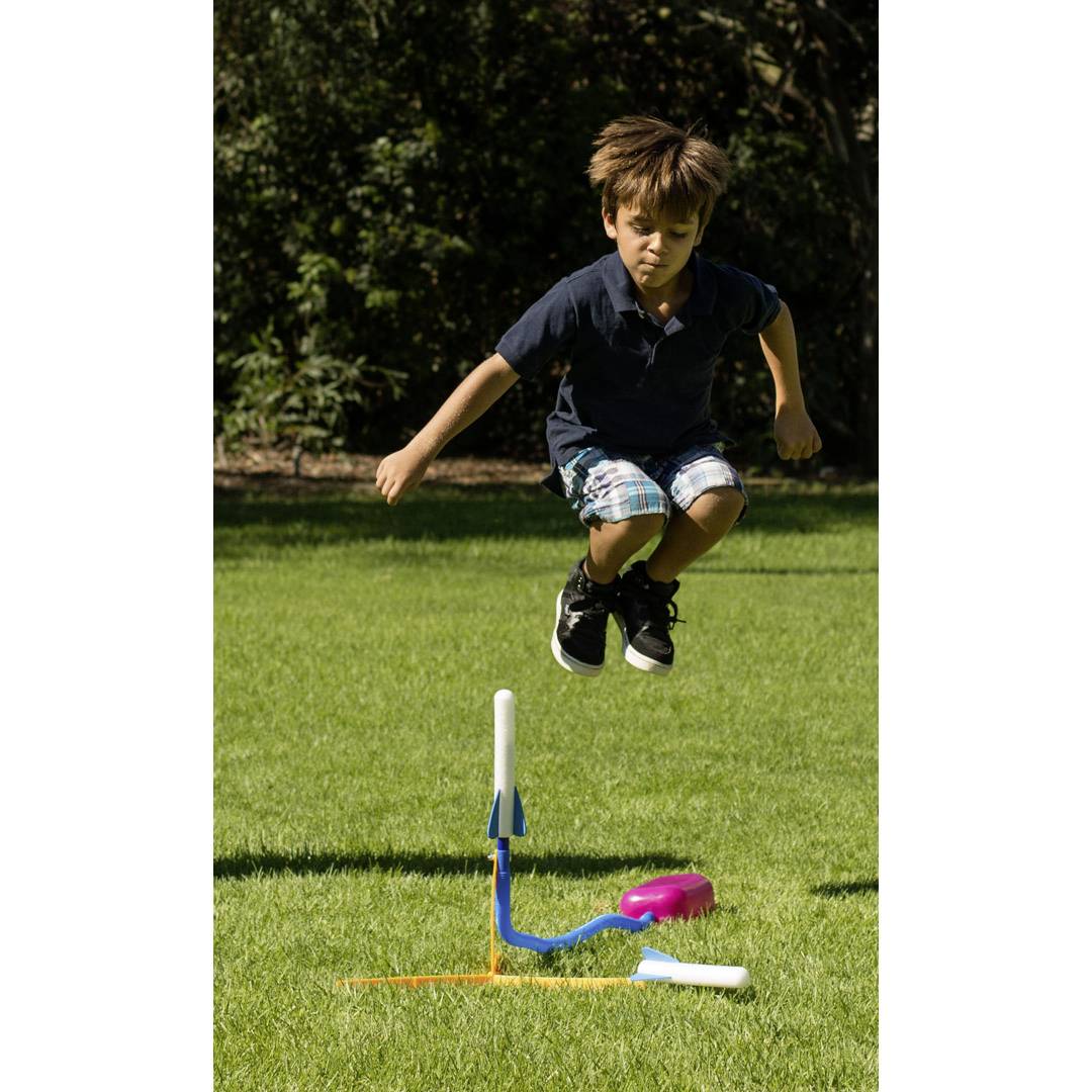 A boy leaps powerfully into the air and lands on a foot pump, which launches a plastic rocket vertically into the sky.