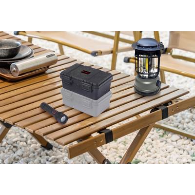 A folding wooden table outdoors with a lantern, a torch, an aluminium container, and a crate placed on top, surrounded by gravel ground.