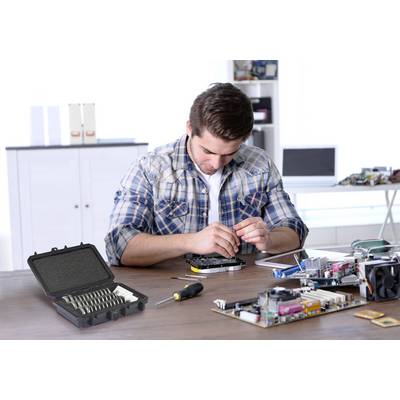 A man is repairing an electronic device at a desk. Tools and electronic components are scattered on the table. Shelves and accessories can be seen in the background.