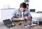A man is repairing an electronic device at a desk. Tools and electronic components are scattered on the table. Shelves and accessories can be seen in the background.