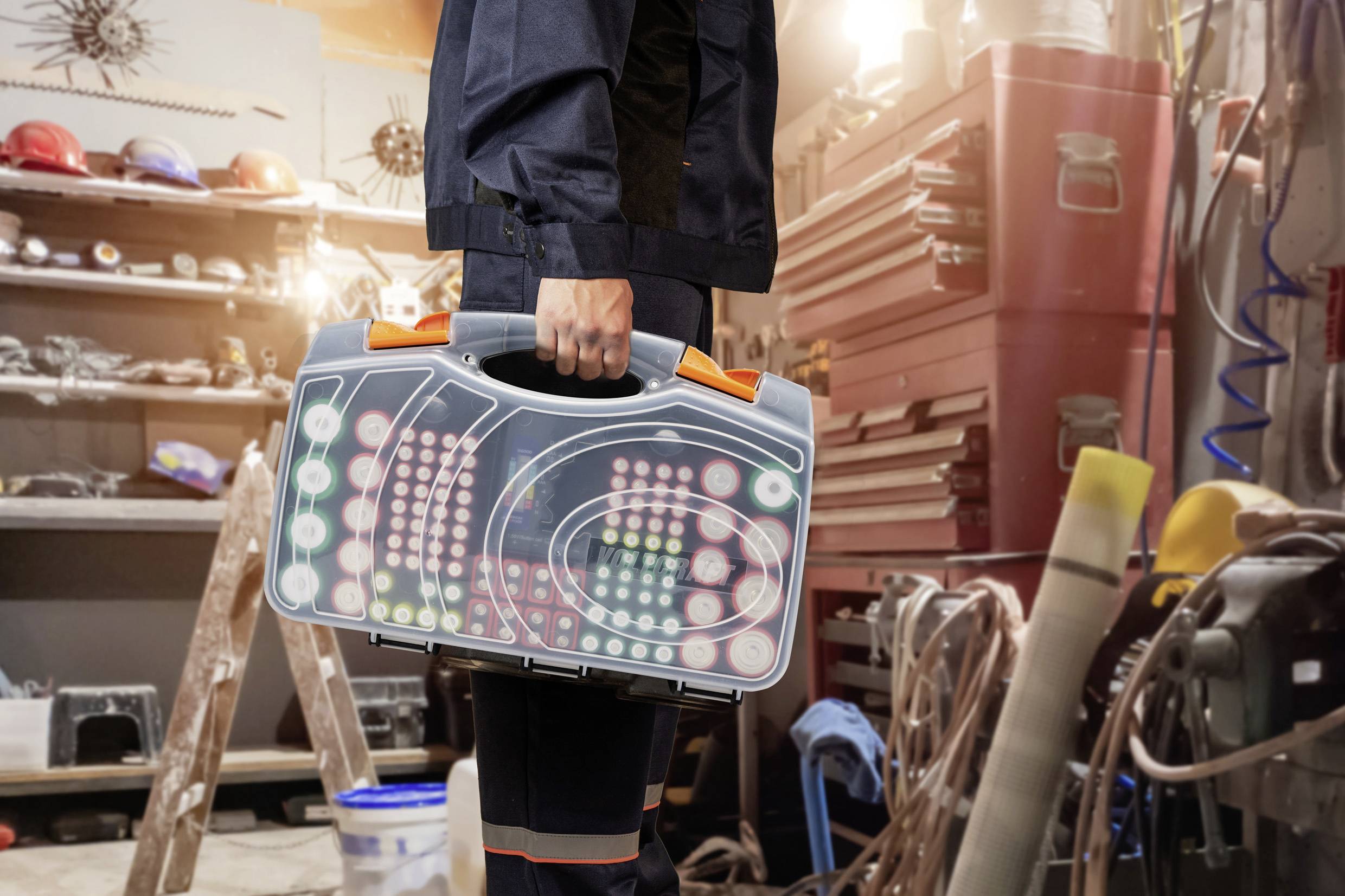 A technician is carrying a case full of colourful buttons and indicators in a workshop. Shelves and tools are visible in the background.