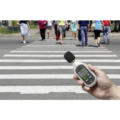 A hand is holding a sound level meter displaying 95.2 decibels. In the background, people are crossing a zebra crossing.