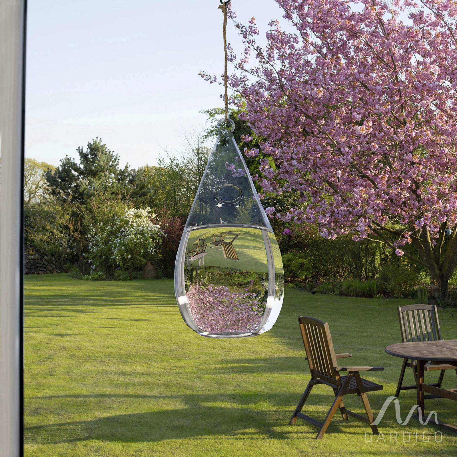A droplet-like wind chime reflects a blossoming cherry tree and stands in a green garden with a pear-shaped chair.