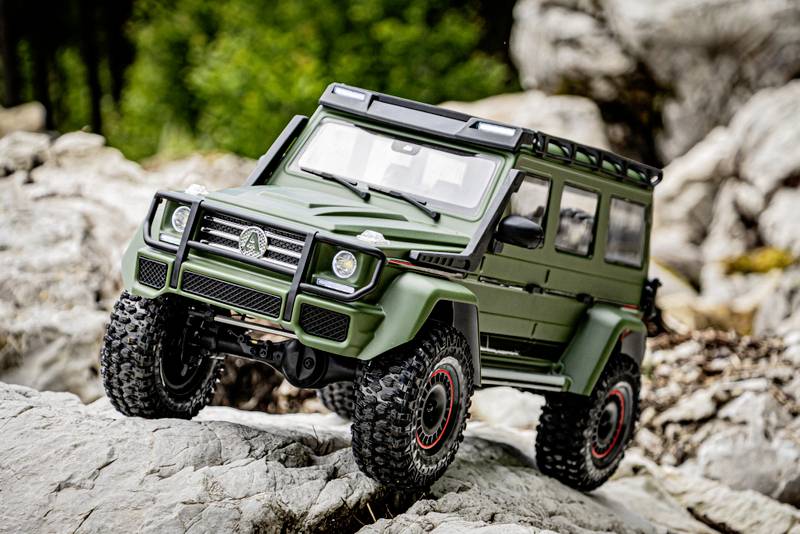 A green toy off-road vehicle positioned on rocky terrain, featuring a typical SUV style with large tyres, with natural landscape in the background.