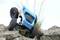 A remote-controlled car with a blue bodywork drives over uneven terrain and rocks, with grass visible in the background.