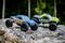 Two remote-controlled model cars are driving over rocky terrain in the woods. One car is blue, the other green. Both have large tyres.