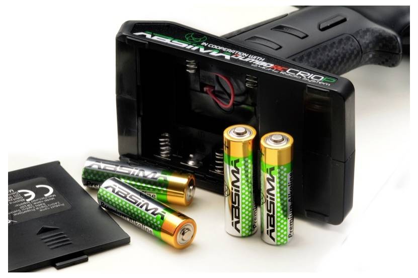 A remote control with its battery compartment cover removed, three AA batteries, and a button cell are lying side by side on a table.