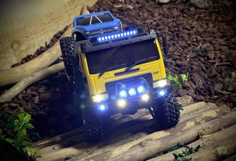 Two remote-controlled off-road vehicles, a yellow one in front and a blue one behind, are driving along a woody path through a miniature landscape.
