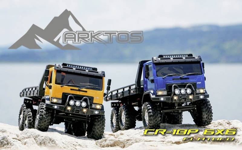 Two remotely controlled, all-terrain lorry models, one orange, one blue, are standing on rocks with the sea in the background.