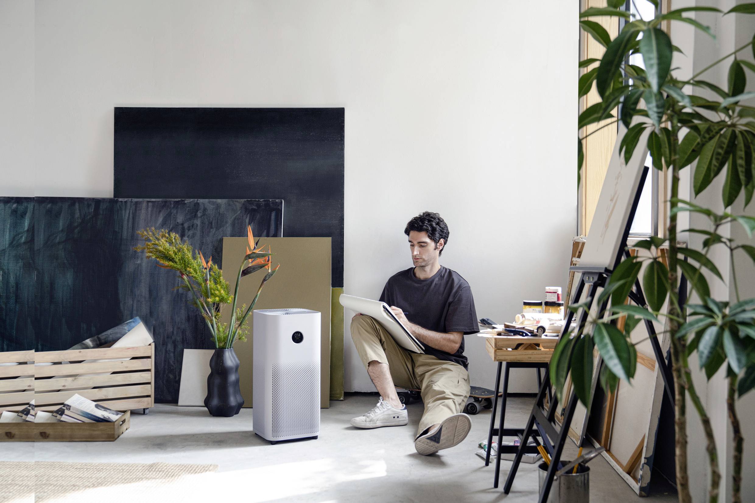 A man is sitting on the floor of a bright room, painting on a pad. A air purifier stands next to him, and there are plants and canvases around.