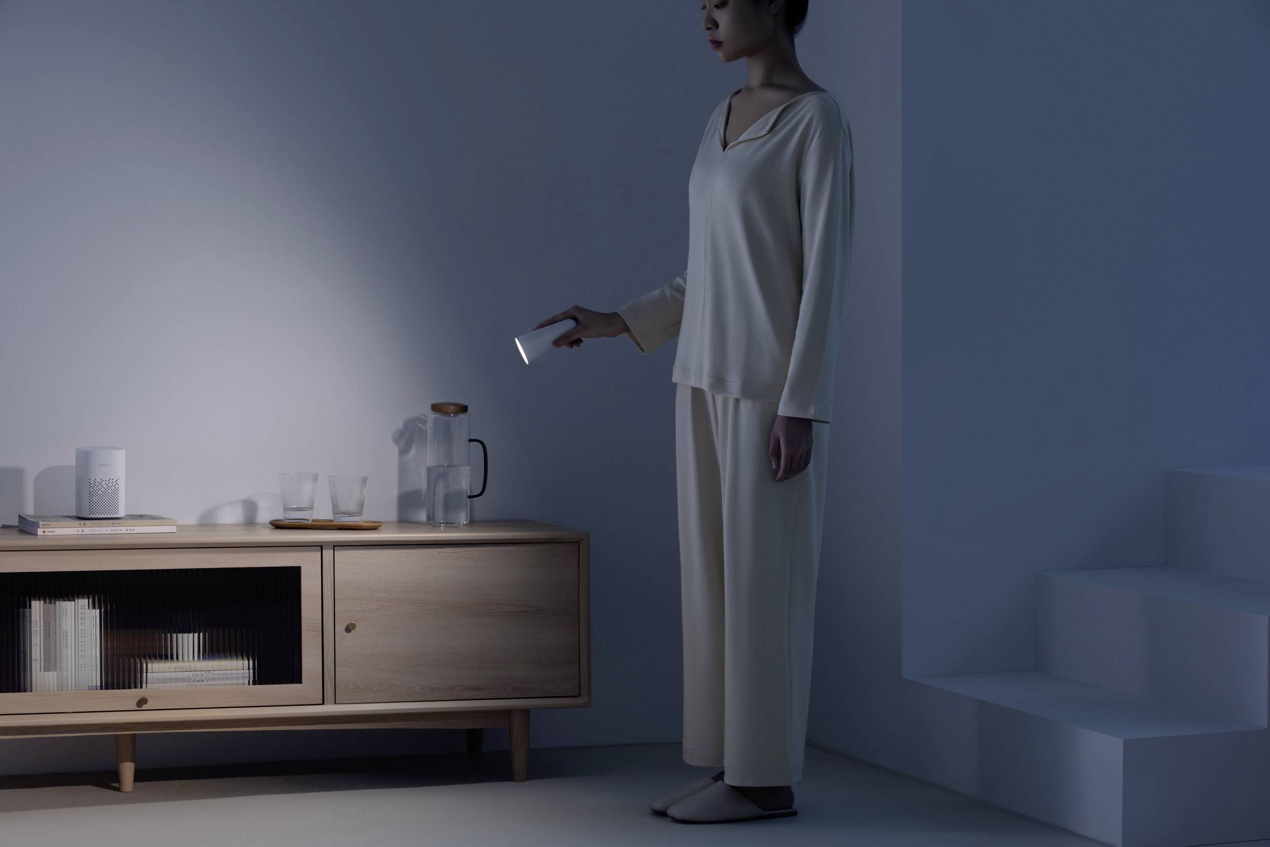 A person in pyjamas is shining a torch in a dimmed room onto a mug and a water jug.
