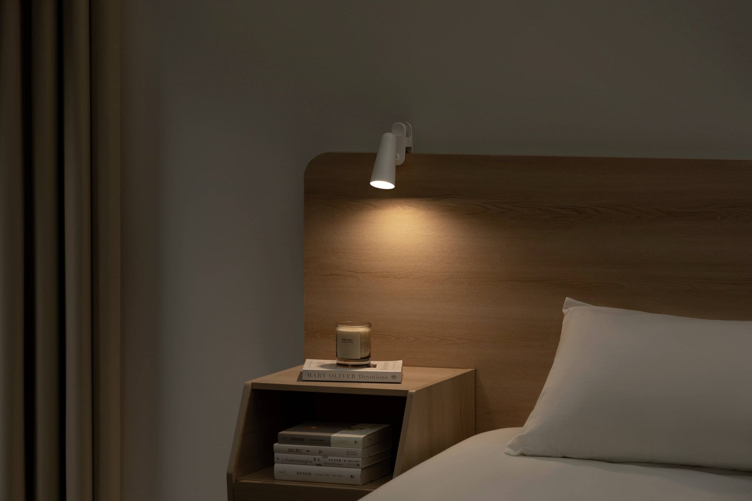 A bedside table with books and a candle stands next to the bed. A wall light above the table casts a warm glow.
