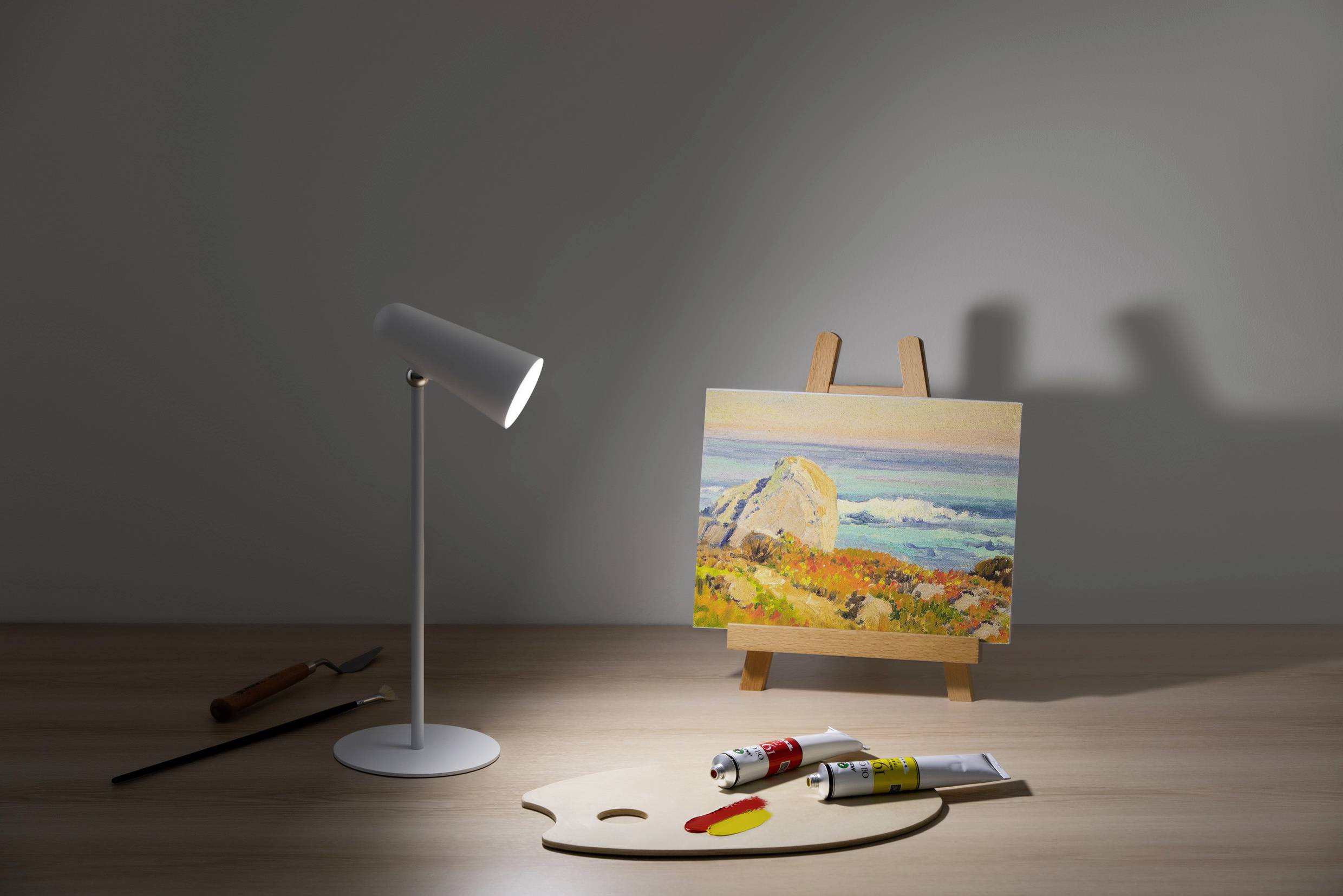 Easel with a landscape painting, beside it a lamp and painting supplies on a table.