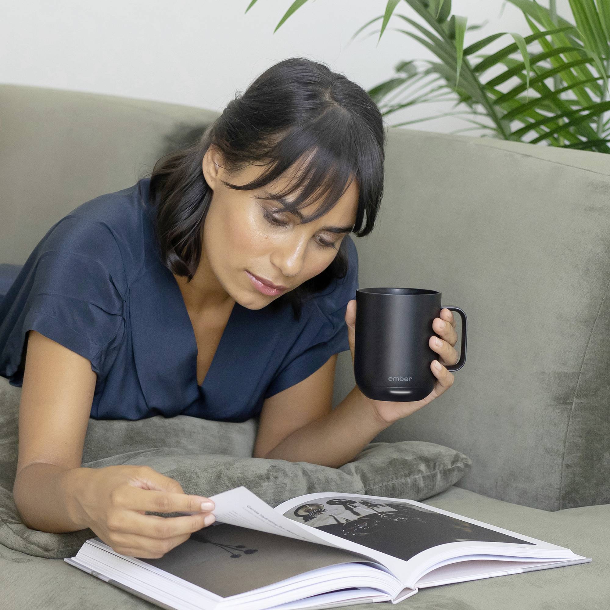 A woman is lying on a sofa reading a book while holding a mug. Plants are visible in the background.