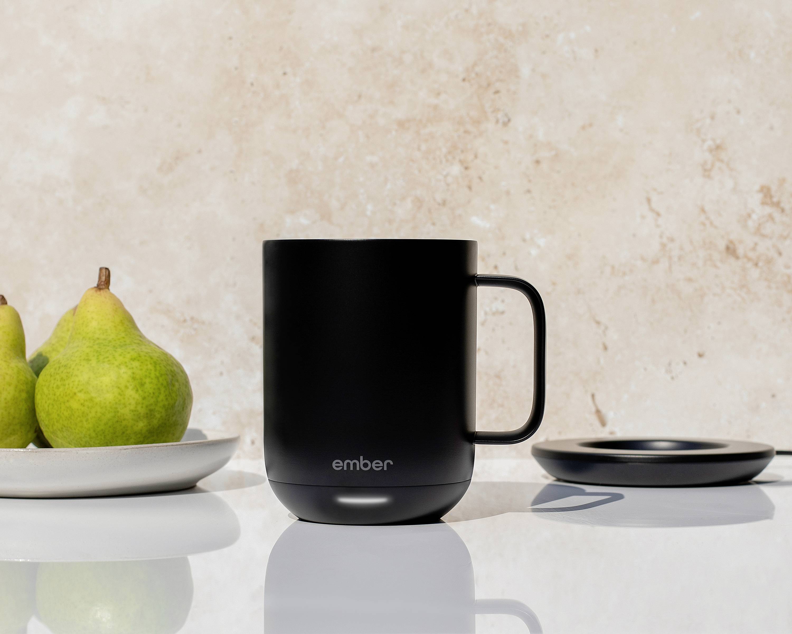 A black mug sits on a table next to two pears on a plate. A beige wall is visible in the background.