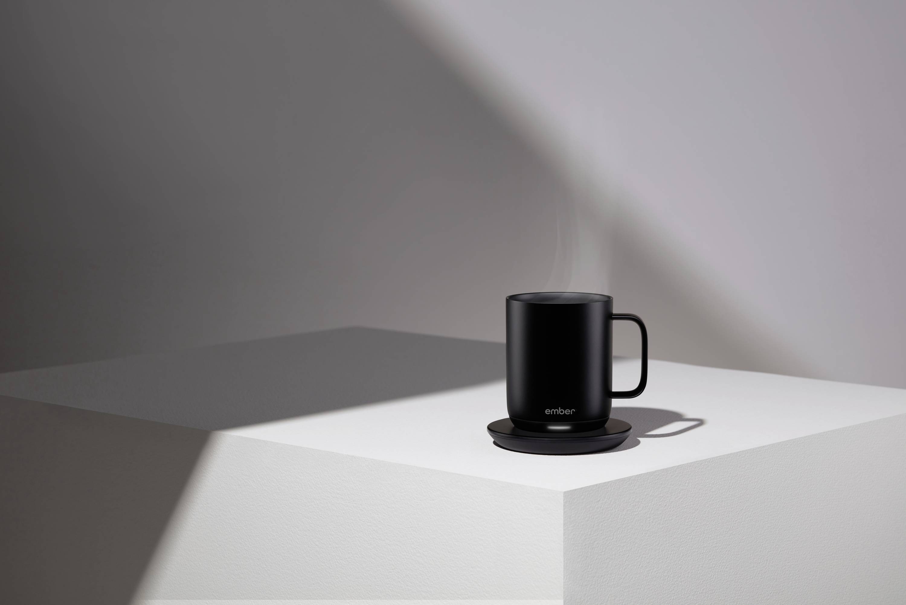 A black thermal mug is standing on a white pedestal. The mug is slightly steaming, indicating a hot liquid inside.