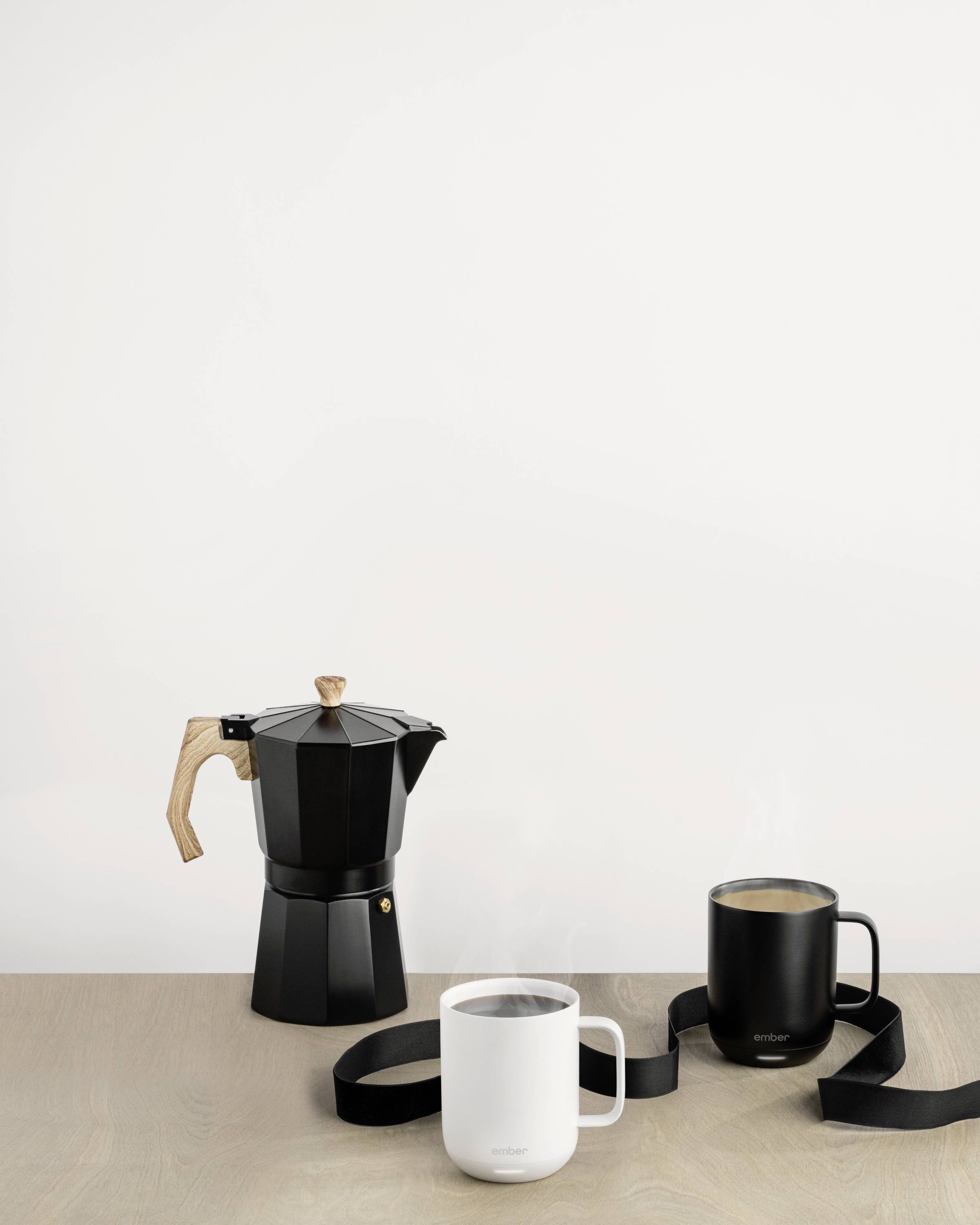 A black espresso pot and two cups (one black, one white) are sitting on a wooden table in front of a white wall.