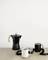 A black espresso pot and two cups (one black, one white) are sitting on a wooden table in front of a white wall.