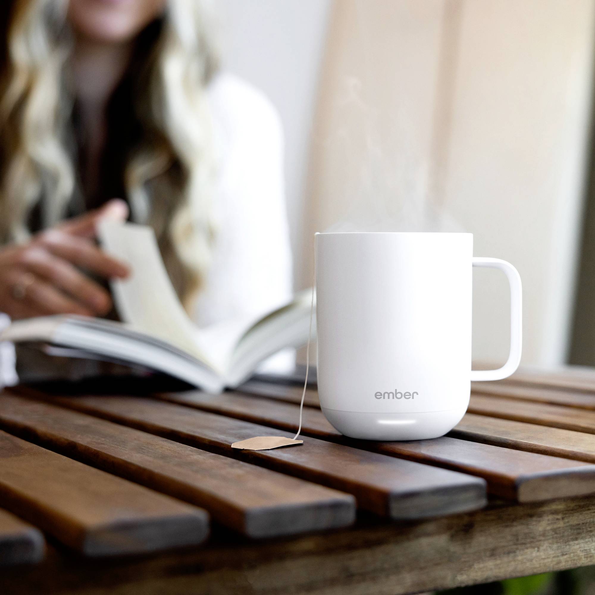 A white cup with steam rising from it sits on a wooden table. In the background, a person is reading a book.