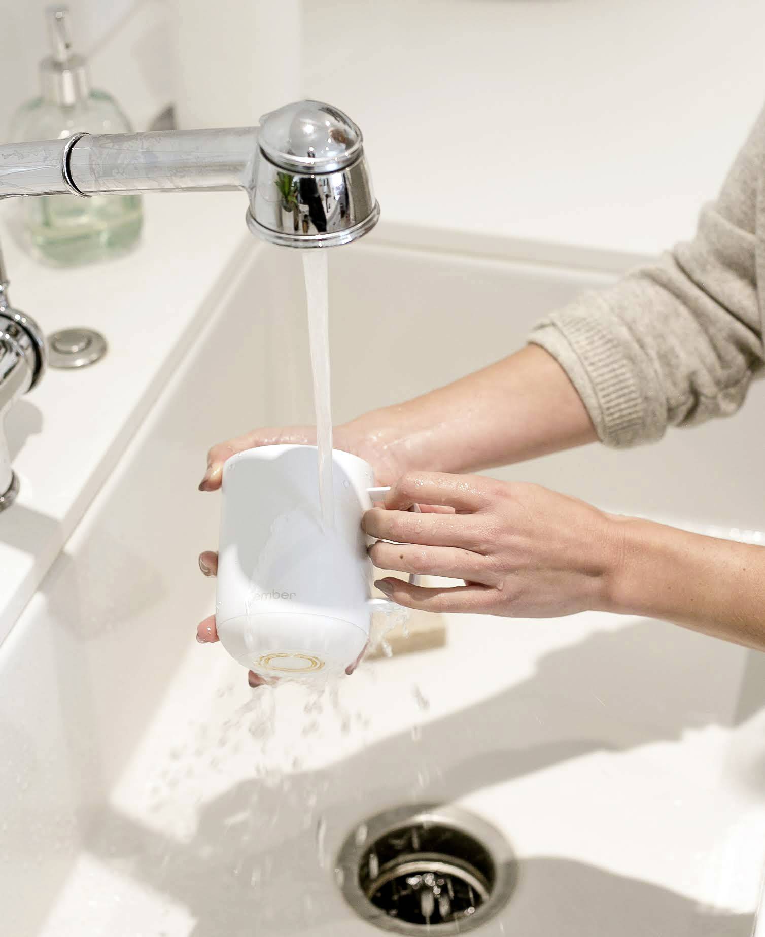 A person is rinsing a white cup under running water in a sink. The right hand is holding the cup, while the left hand is cleaning it.