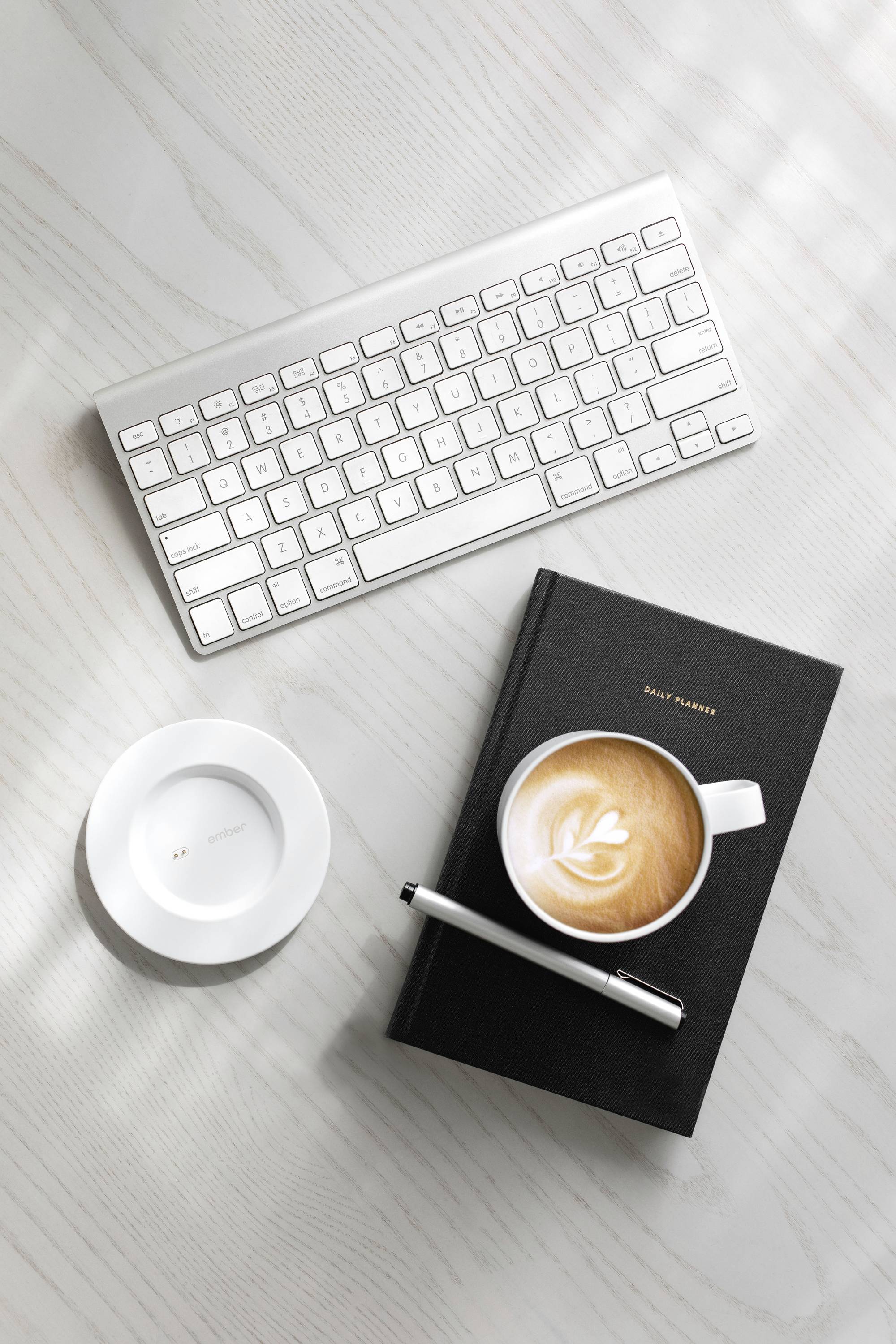 Keyboard, notebook with pen and cup of cappuccino on a desk. Office workspace with drink and writing materials.