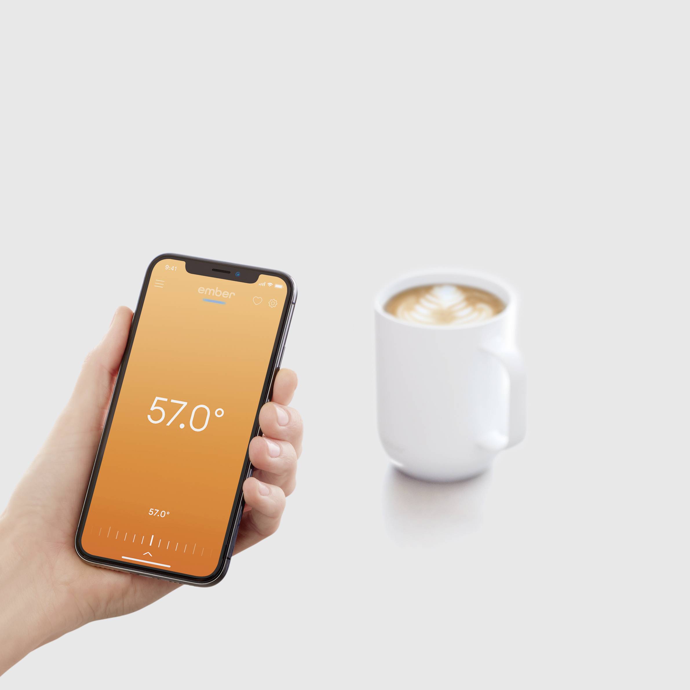 A hand is holding a smartphone displaying a temperature of 57 degrees Celsius. In the background, a coffee cup with latte art is visible.