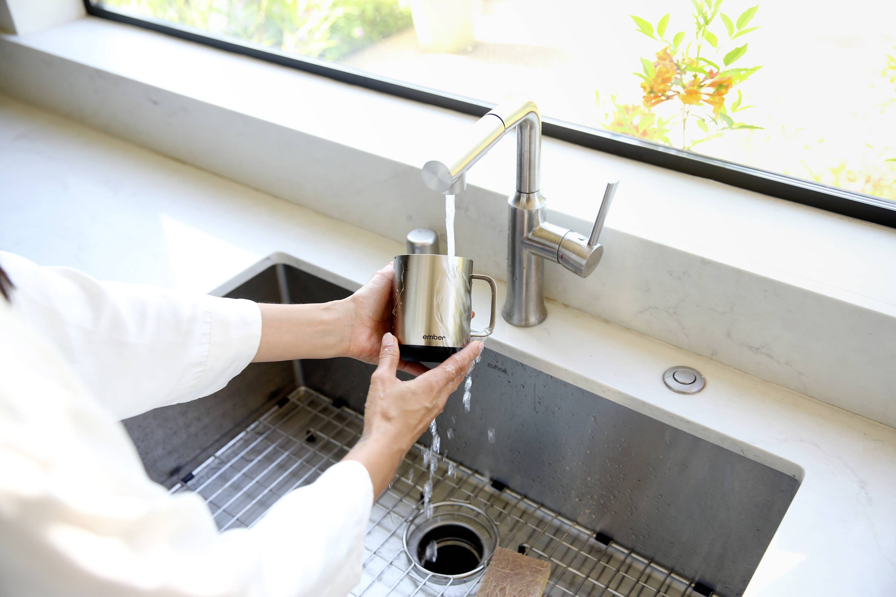 A person fills a metal mug with water from a kitchen tap at the sink. Bright daylight streams through the window behind.