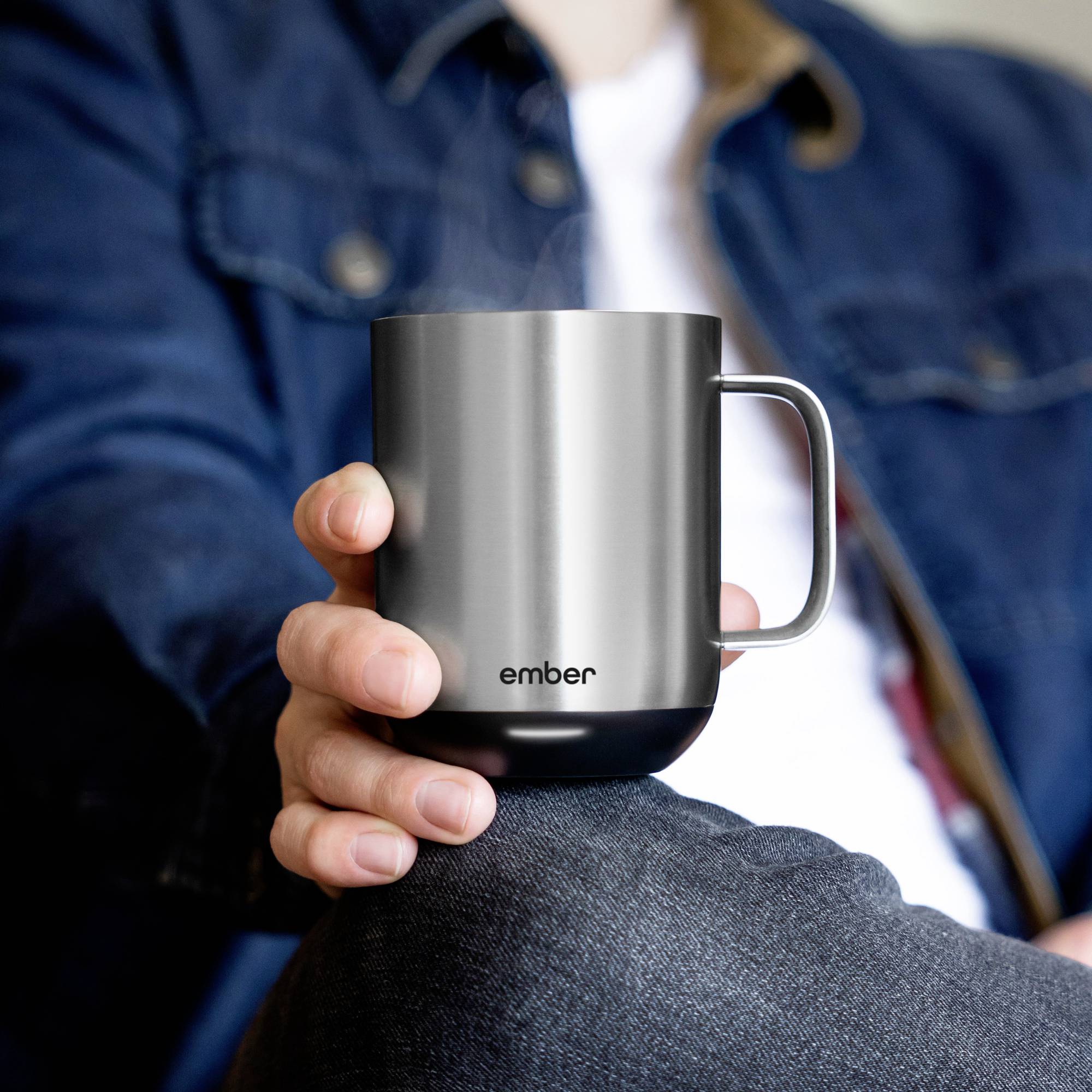 A person is holding a grey mug with the word 'ember' printed on it, with steam rising from it. The person is wearing a navy jacket.