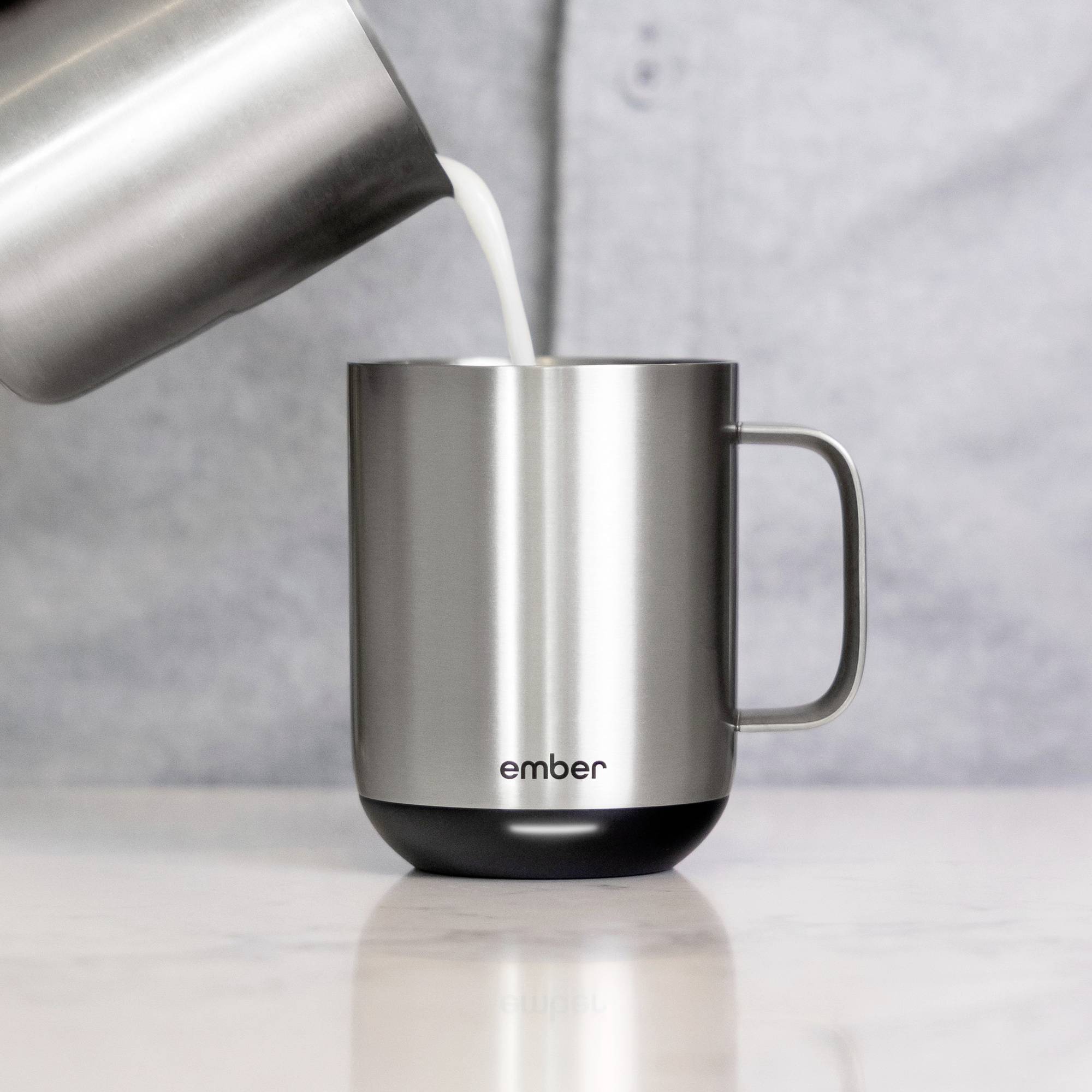 A stainless steel mug with 'ember' printed on it is being filled with milk. The mug is sitting on a grey surface.