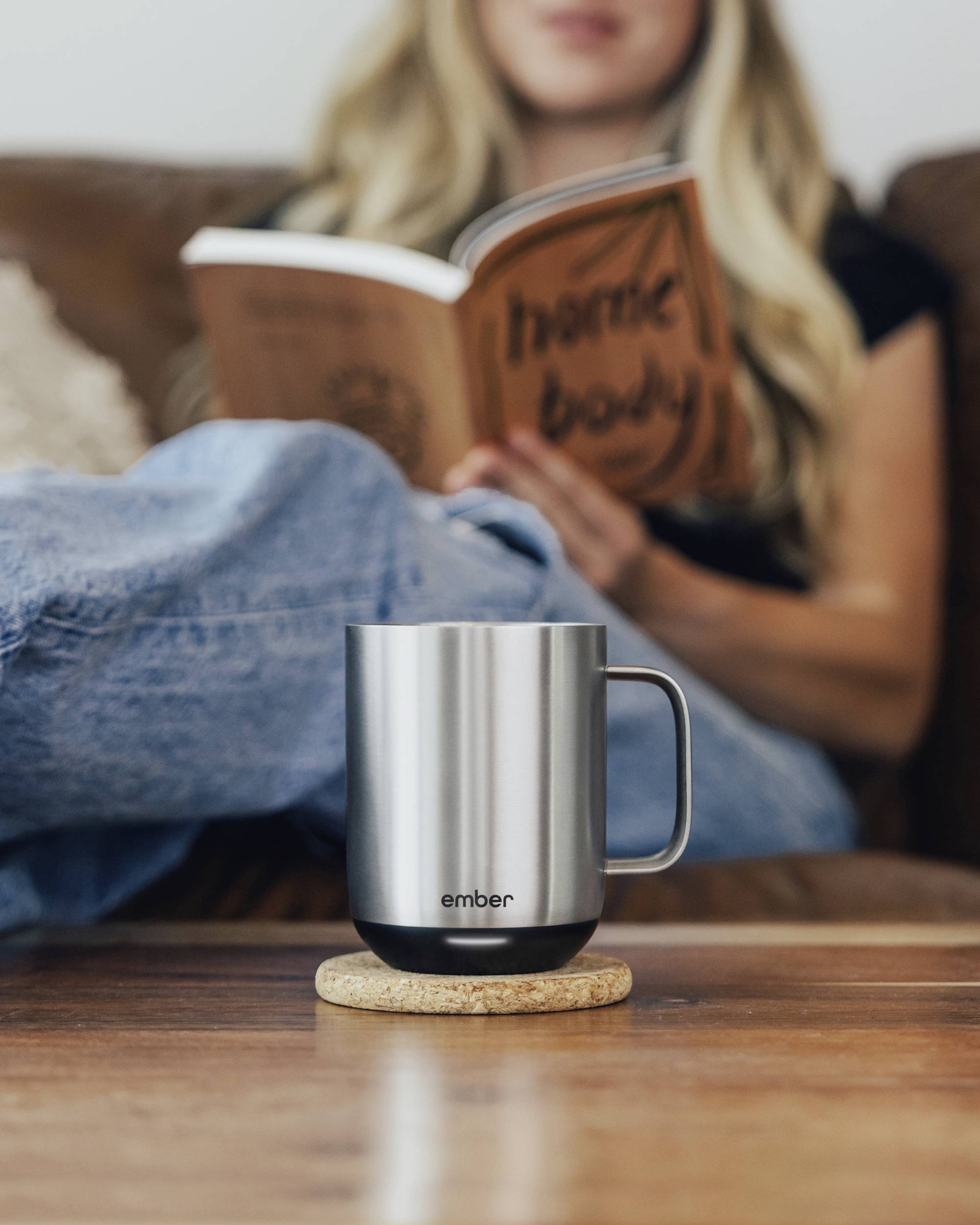 A person is sitting on a sofa reading a book. In the foreground, a silver thermal mug stands on a table.