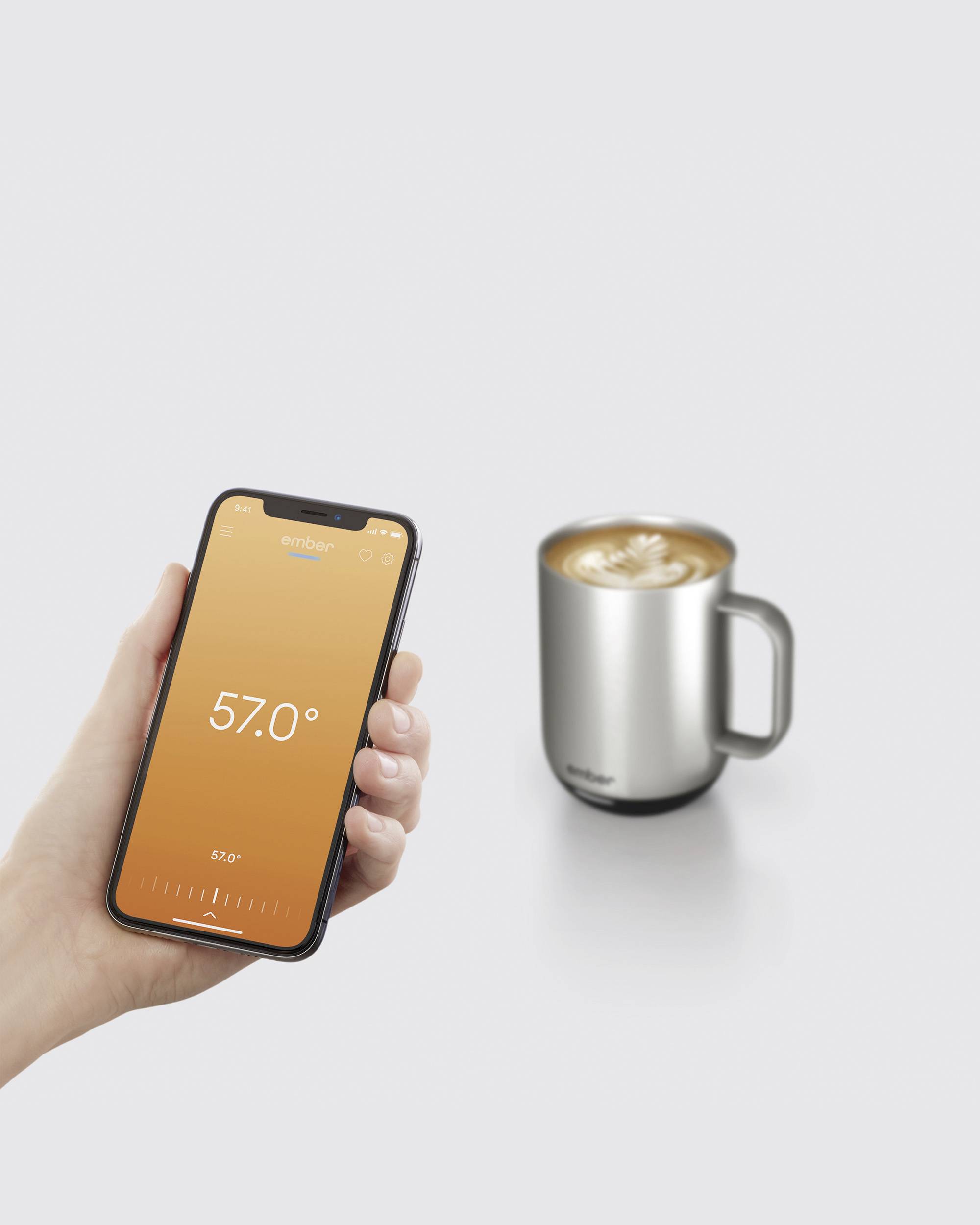 A hand is holding a smartphone displaying a temperature of 57 degrees. Beside it sits a silver cup with a hot drink.