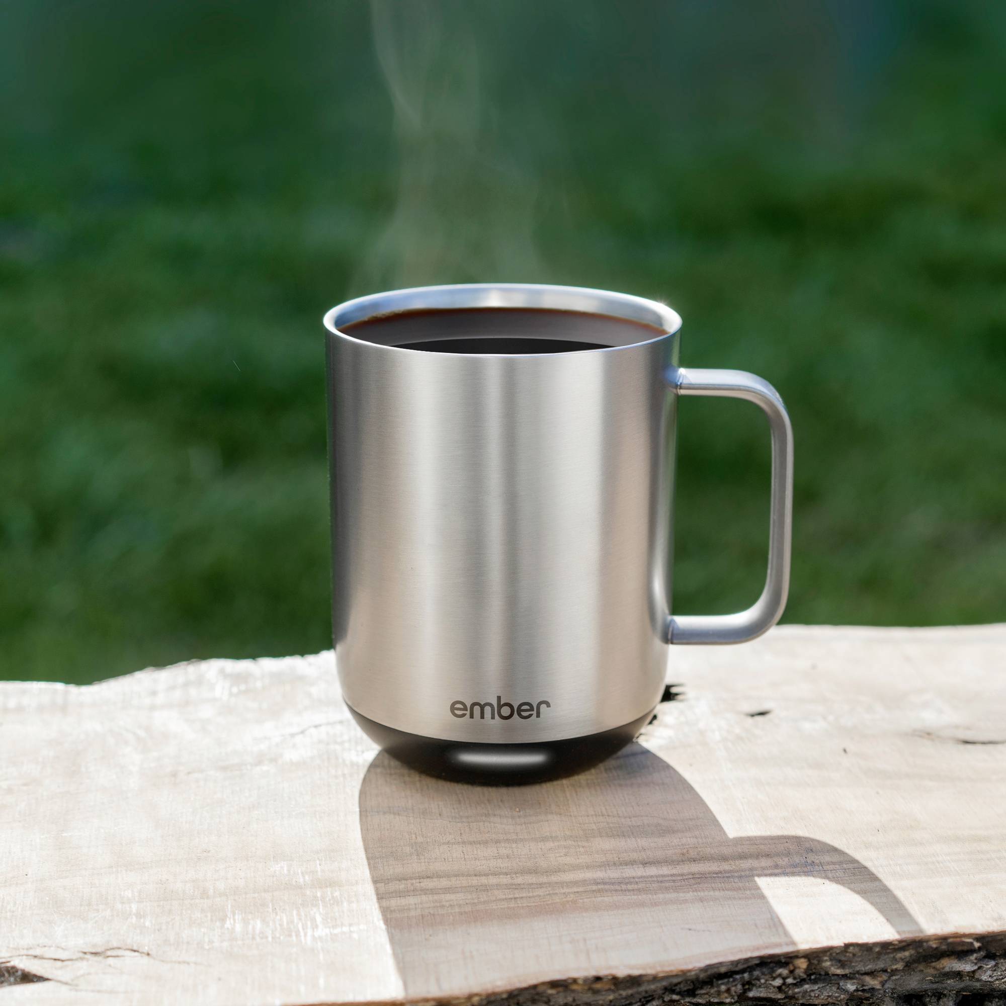 Metal mug with a steaming drink on a wooden table outdoors. The mug has a handle, and the 'ember' brand is depicted on it.