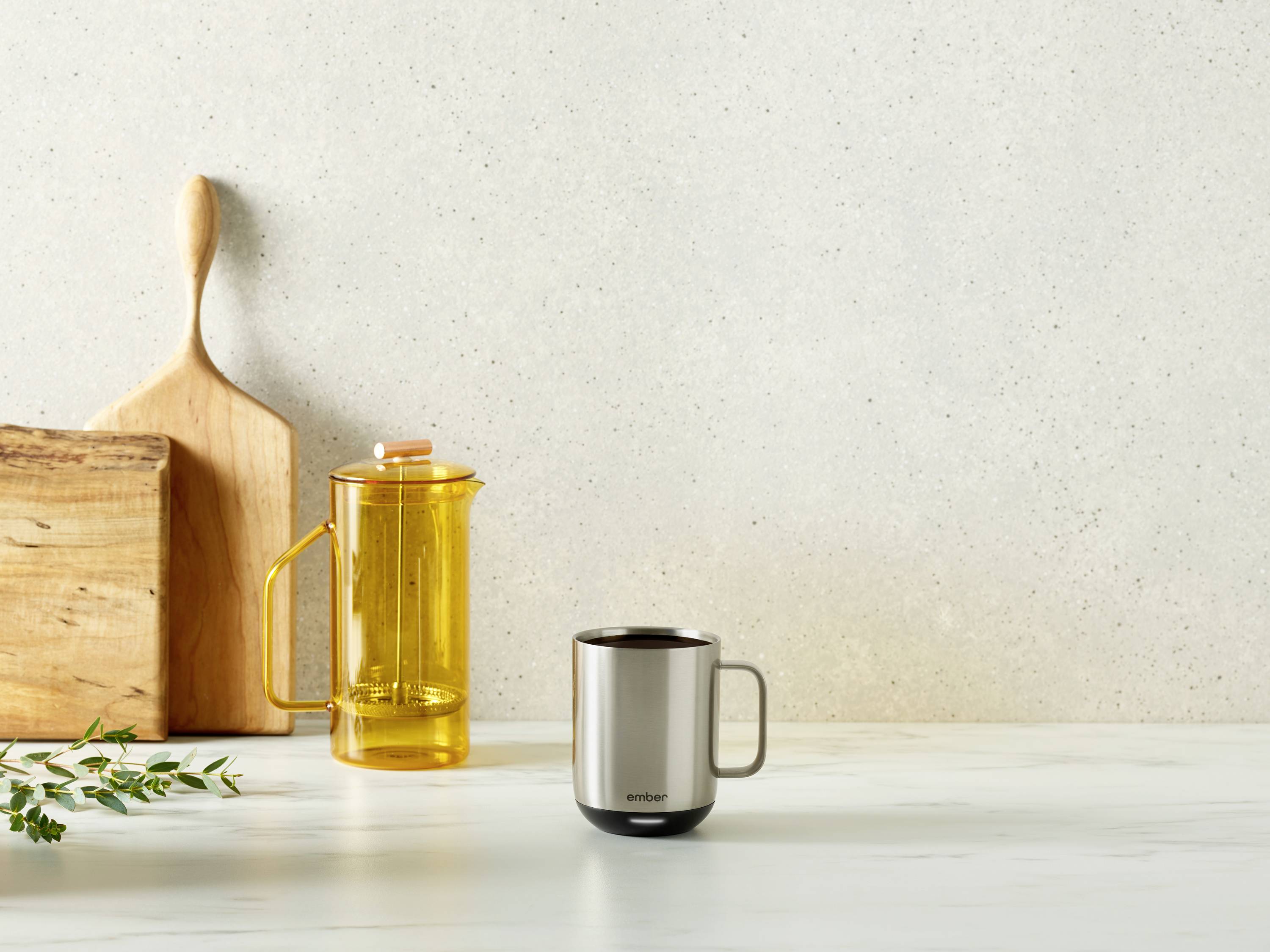 A metal mug on a marble table, next to a yellow teapot and a wooden chopping board against a grey background.