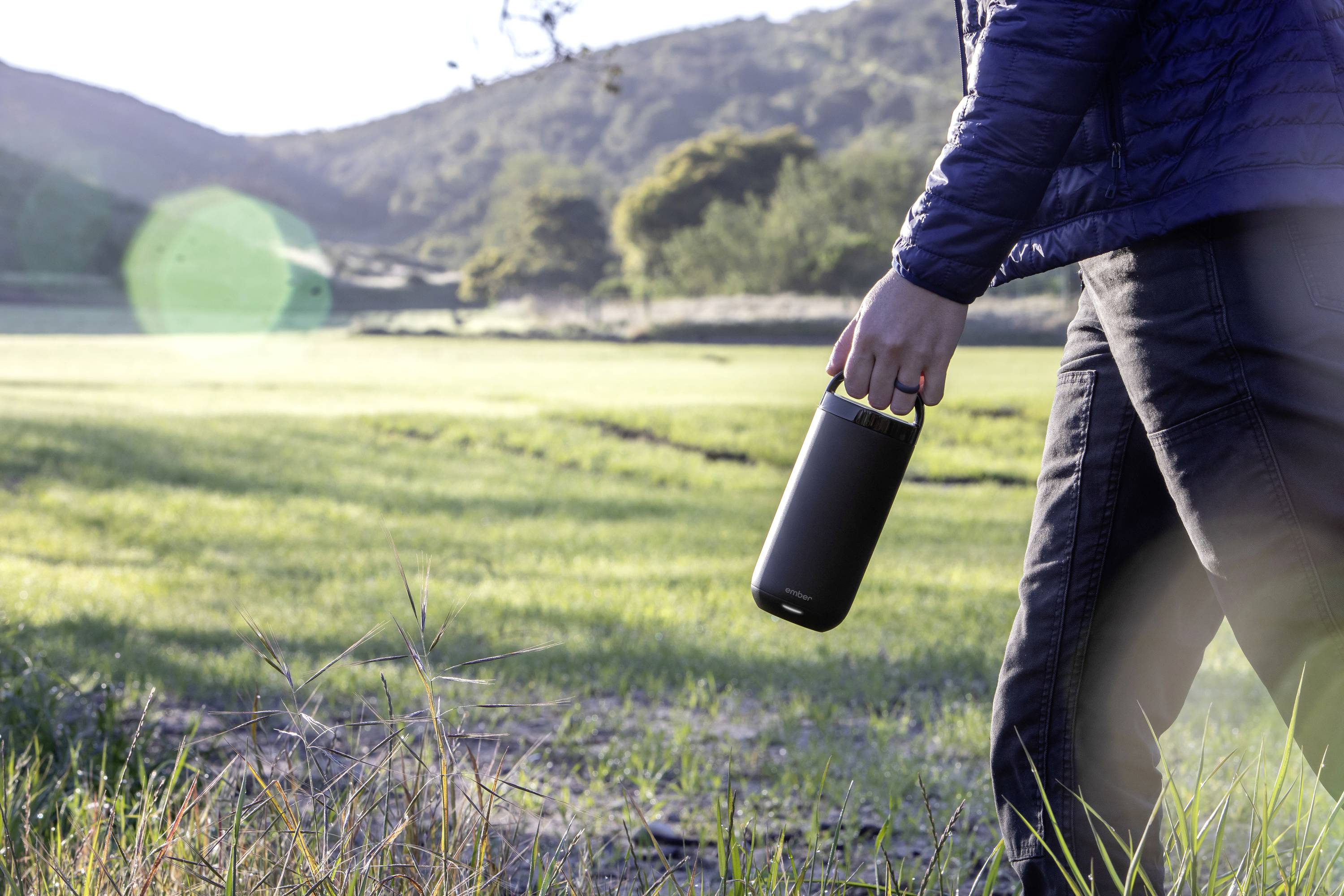 A person is walking through a green meadow, holding a black water bottle. Hills and trees can be seen in the background.