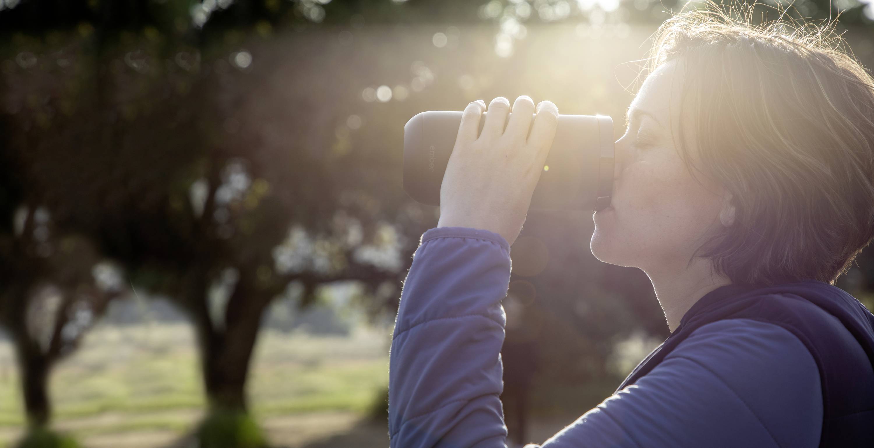 A person in casual clothing is drinking from a thermos outdoors, with sunlight filtering through the trees.