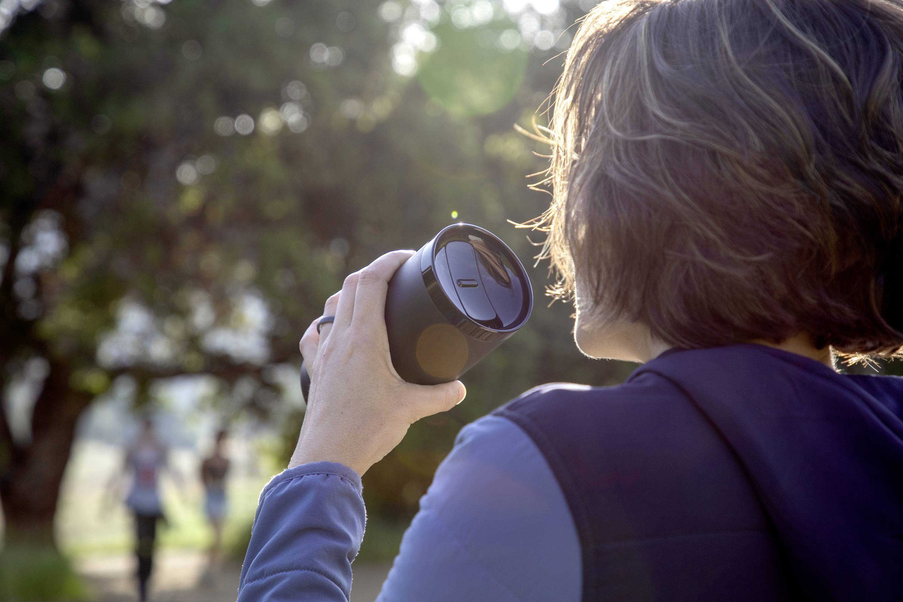 A person with short hair is holding a coffee cup outdoors. Trees and two blurry silhouettes are visible in the background.