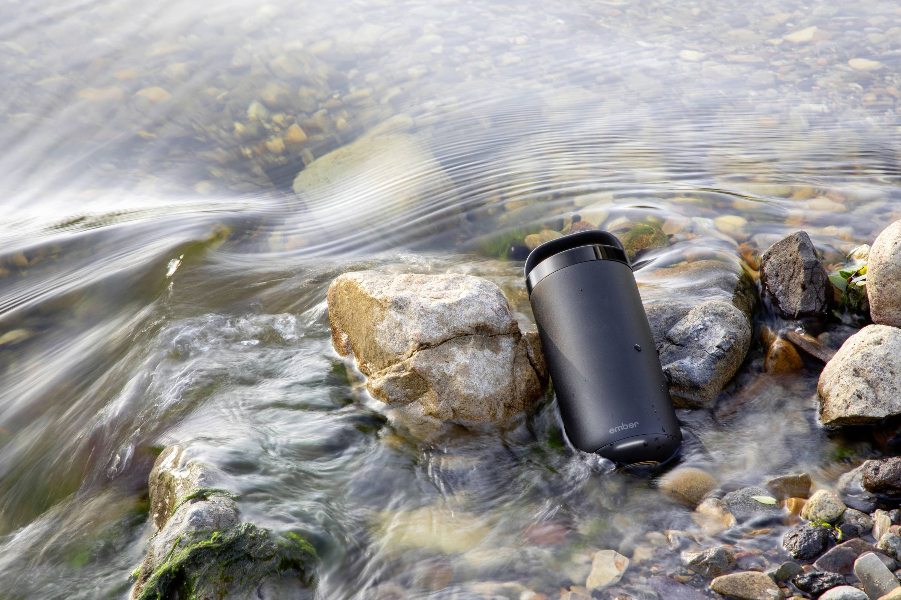 A black thermal mug lies in flowing water between stones at the water's edge.