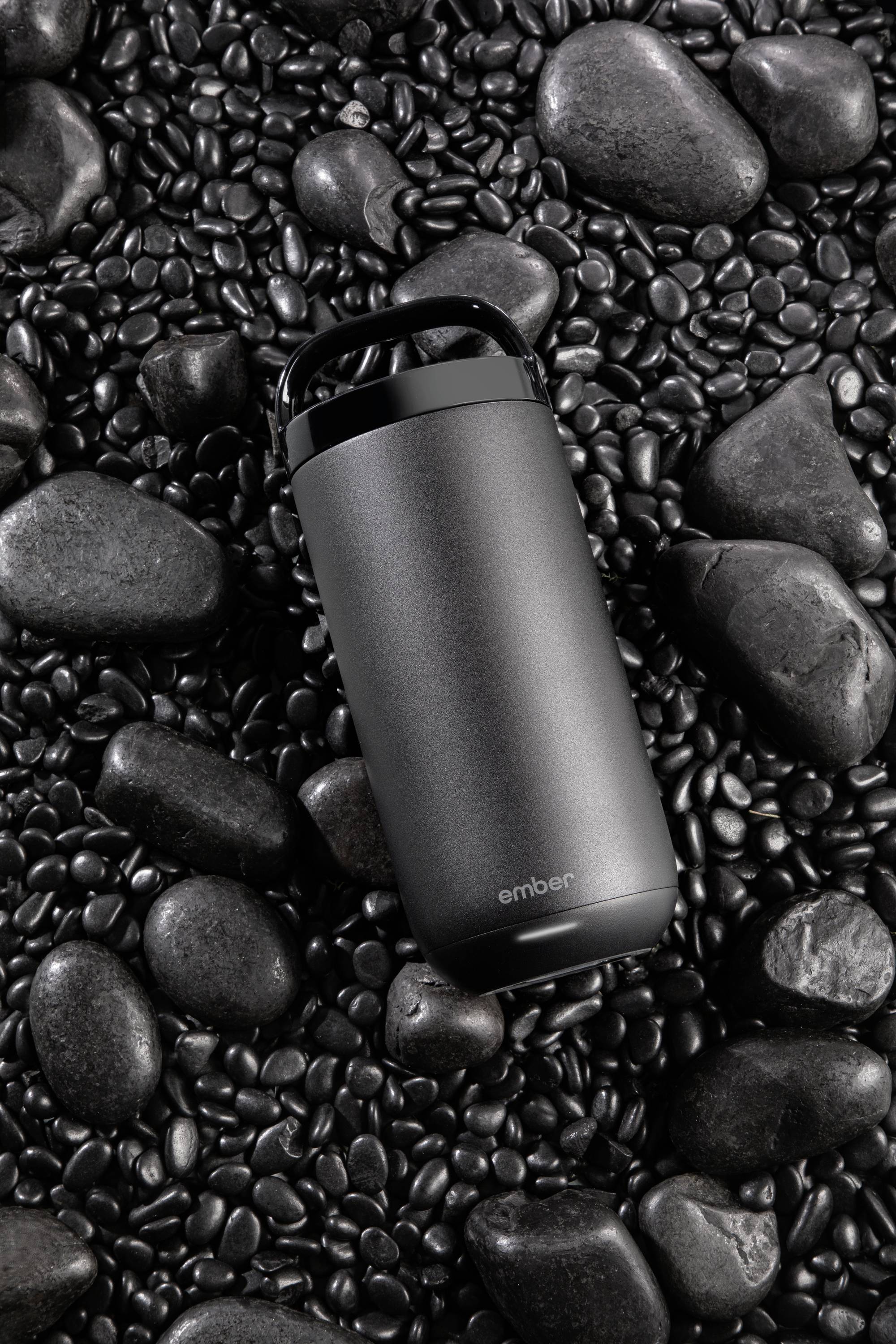 A black thermal mug rests on smooth, dark stones.