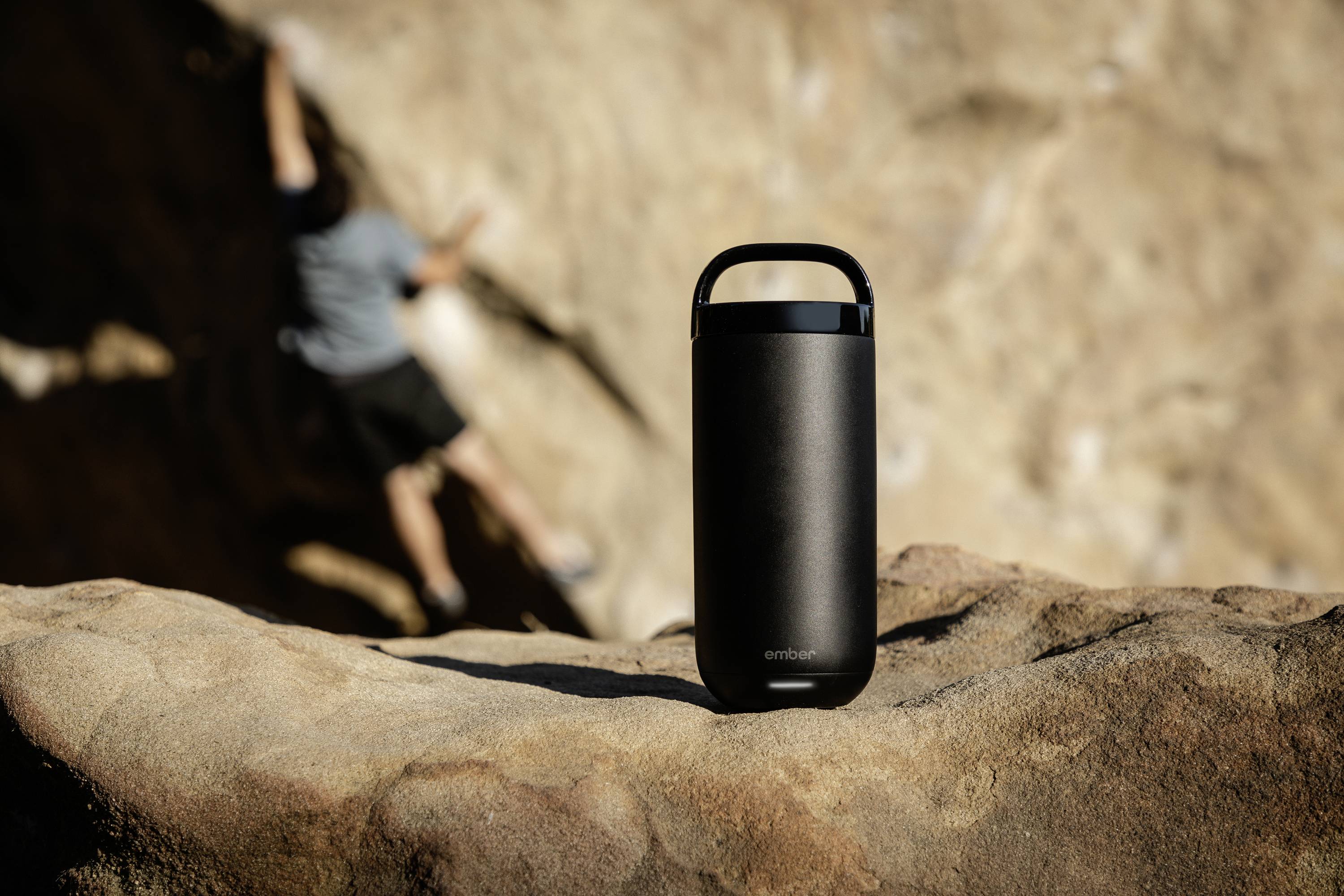 A black portable speaker sits on a rock in the foreground. In the background, a person is climbing a rock face.